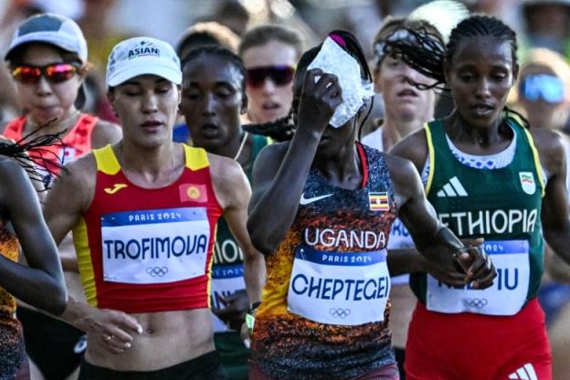 A woman marathon runner, with a bib on her chest bearing the name Cheptegei, holds a bag of ice against her face as she runs among a crowd of other women.